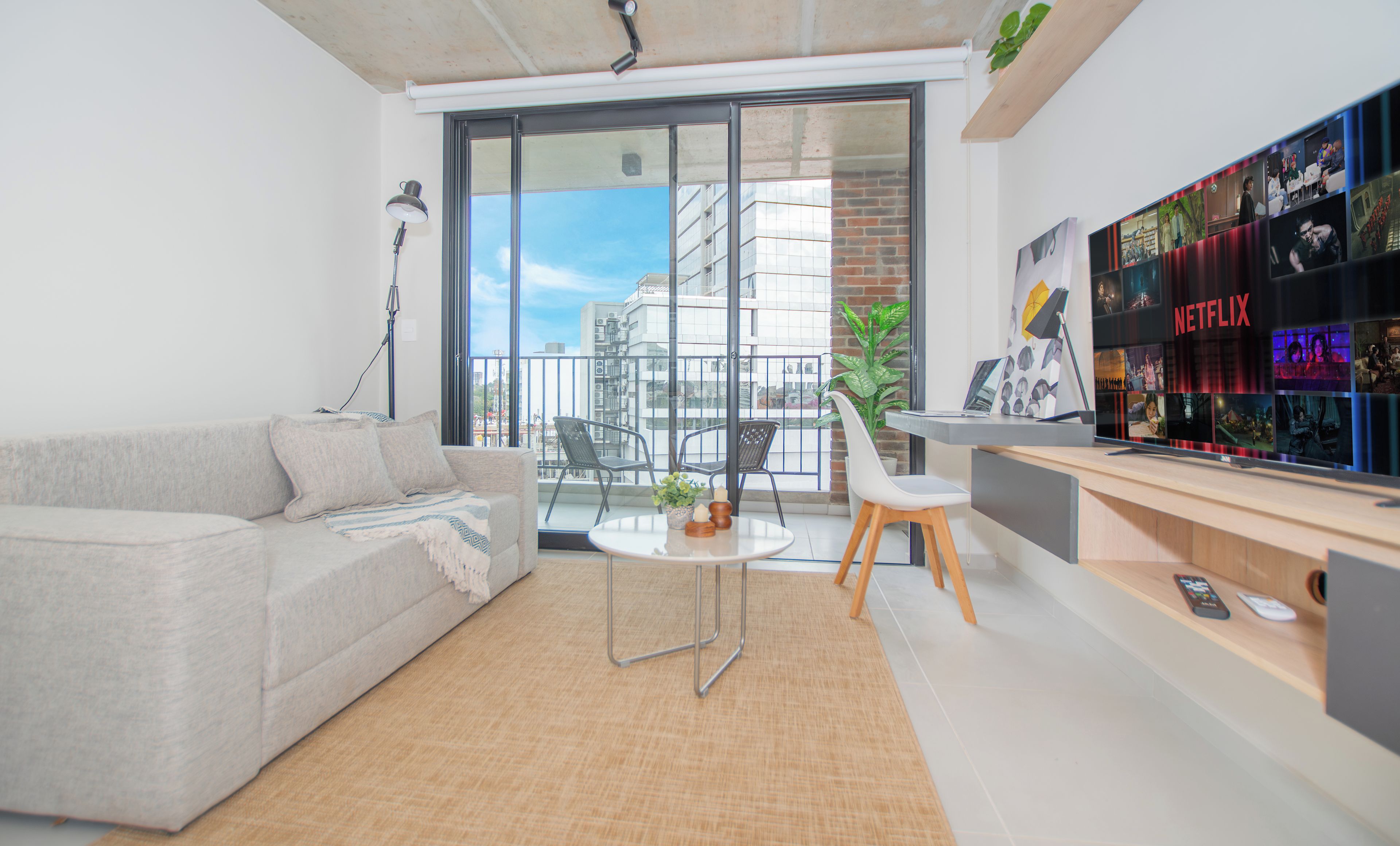 A modern living room with a sofa, a small round table, and a desk with a computer. There is a large window with a view of buildings and a balcony.