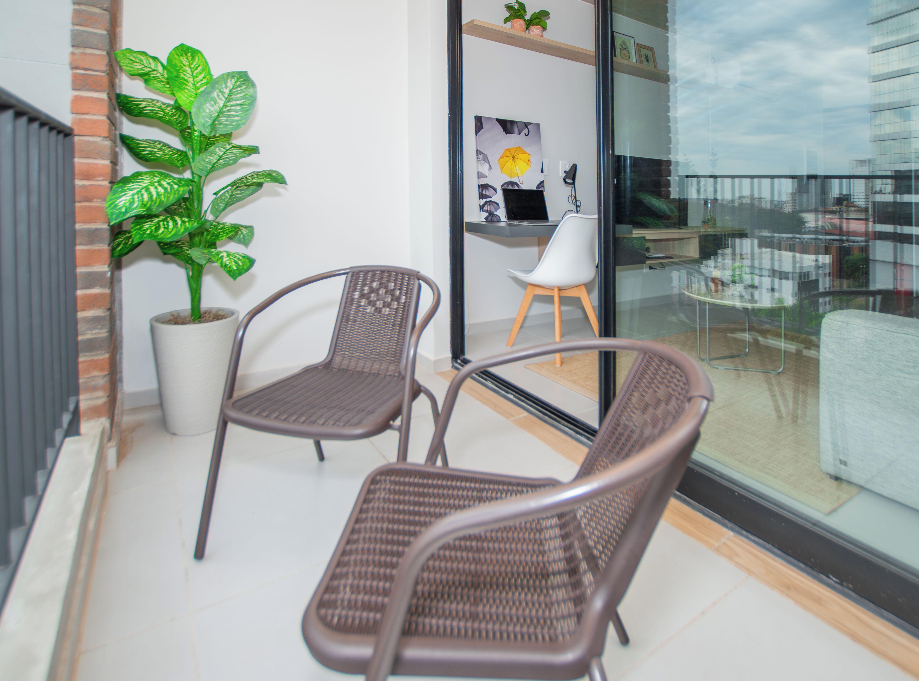 A balcony with two wicker chairs, a potted plant, and a glass door leading to an indoor area with a white chair and a framed picture.