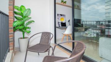 A balcony with two wicker chairs, a potted plant, and a glass door leading to an indoor area with a white chair and a framed picture.