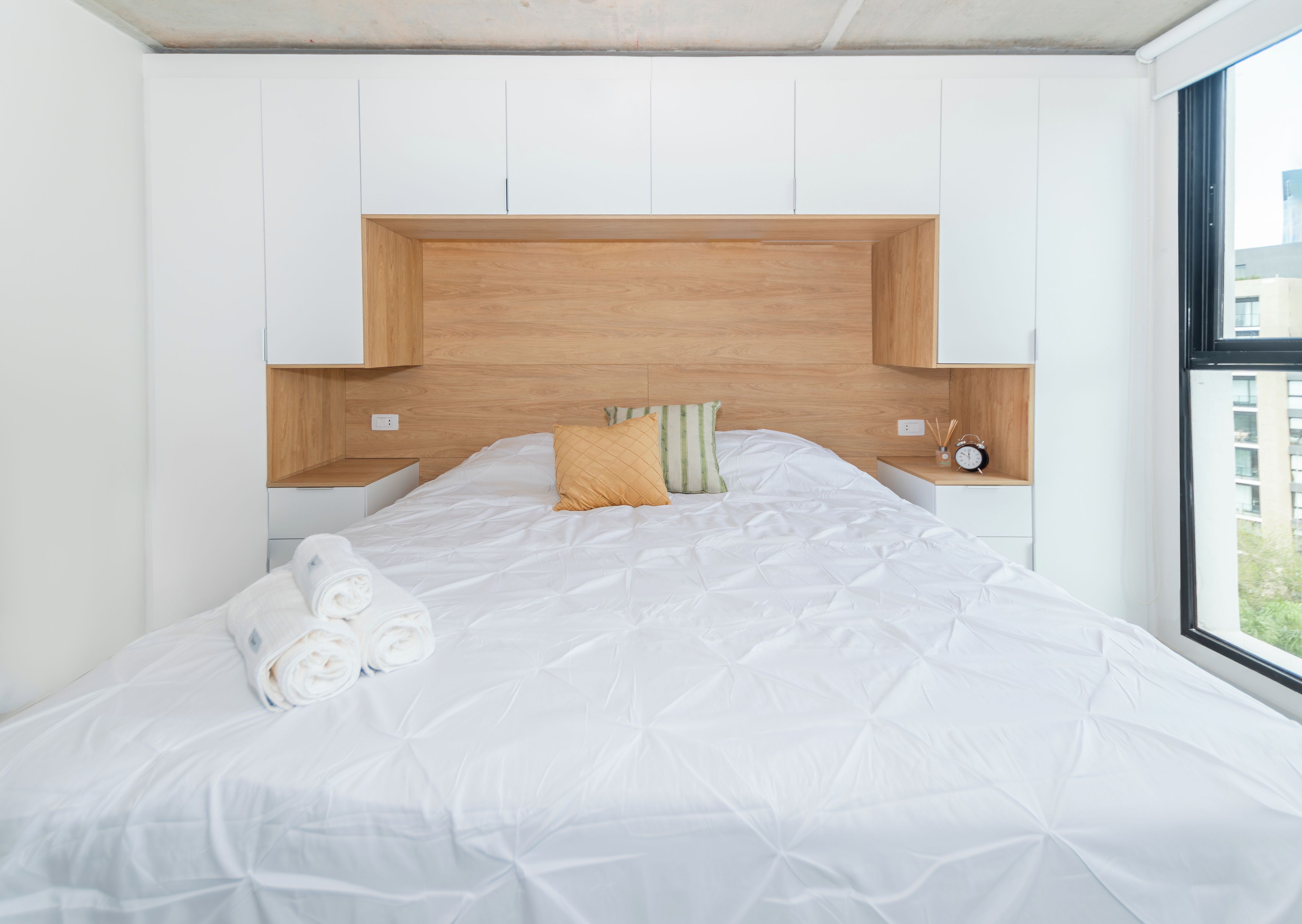 A modern bedroom with a large bed, white bedding, a wooden headboard, and a view of buildings outside the window.
