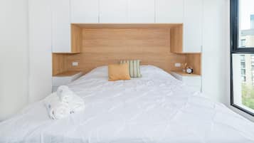 A modern bedroom with a large bed, white bedding, a wooden headboard, and a view of buildings outside the window.