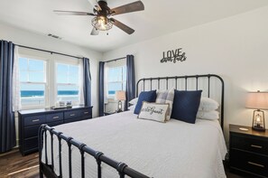 Ein Schlafzimmer mit Bett, Deckenventilator, Nachttisch, Lampe, einem Fenster mit Vorhängen und einem Wandschild.