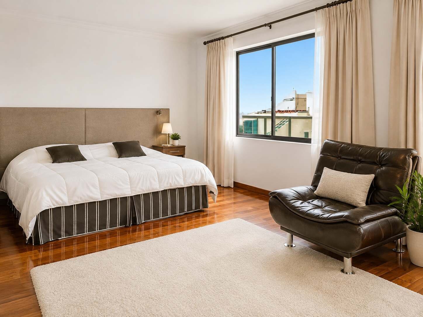 A bedroom with a bed, a nightstand, a window with curtains, a leather armchair, and a potted plant.