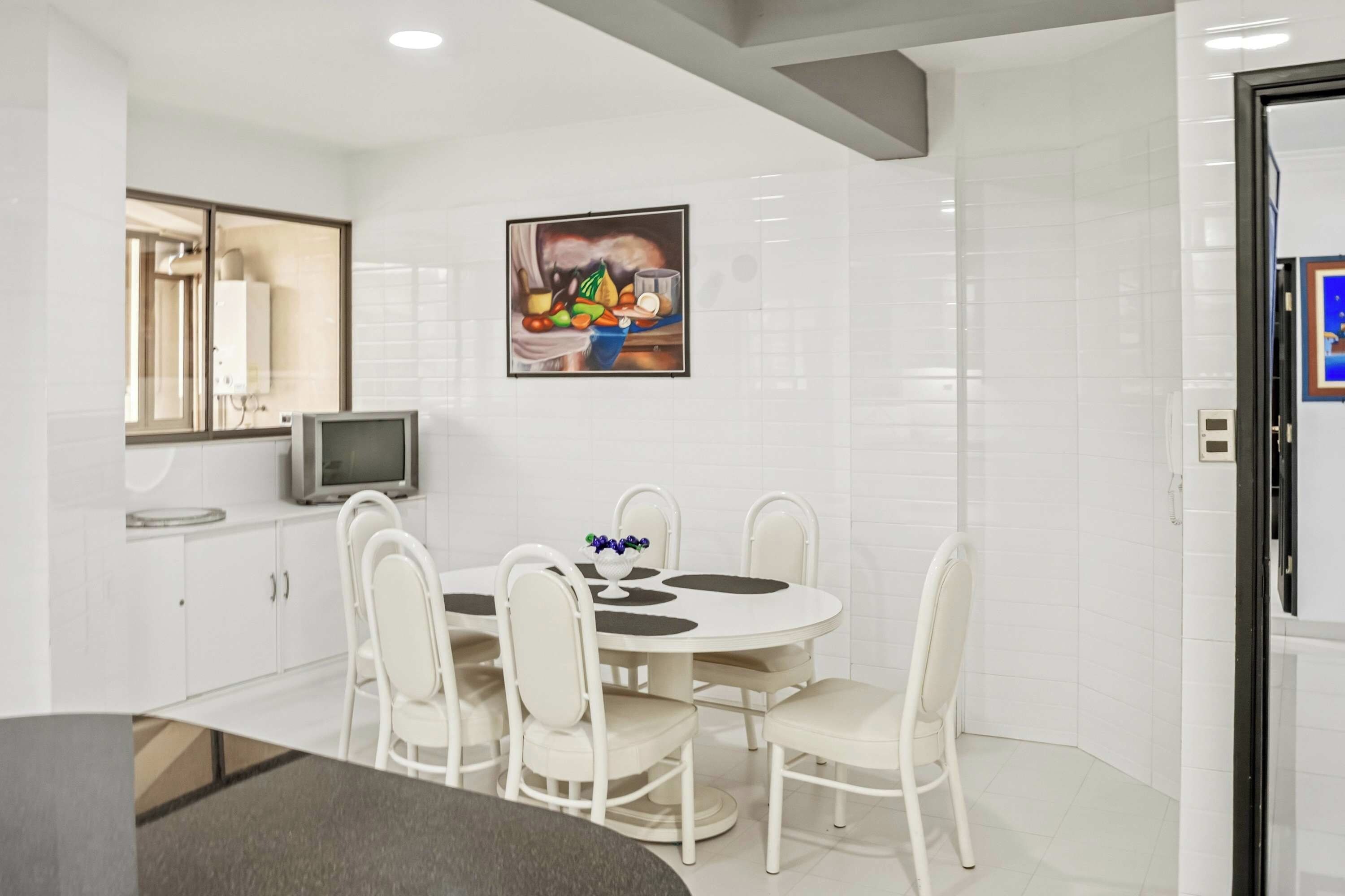 A modern kitchen with white cabinetry, a dining table with chairs, a flat-screen TV, and a painting on the wall.