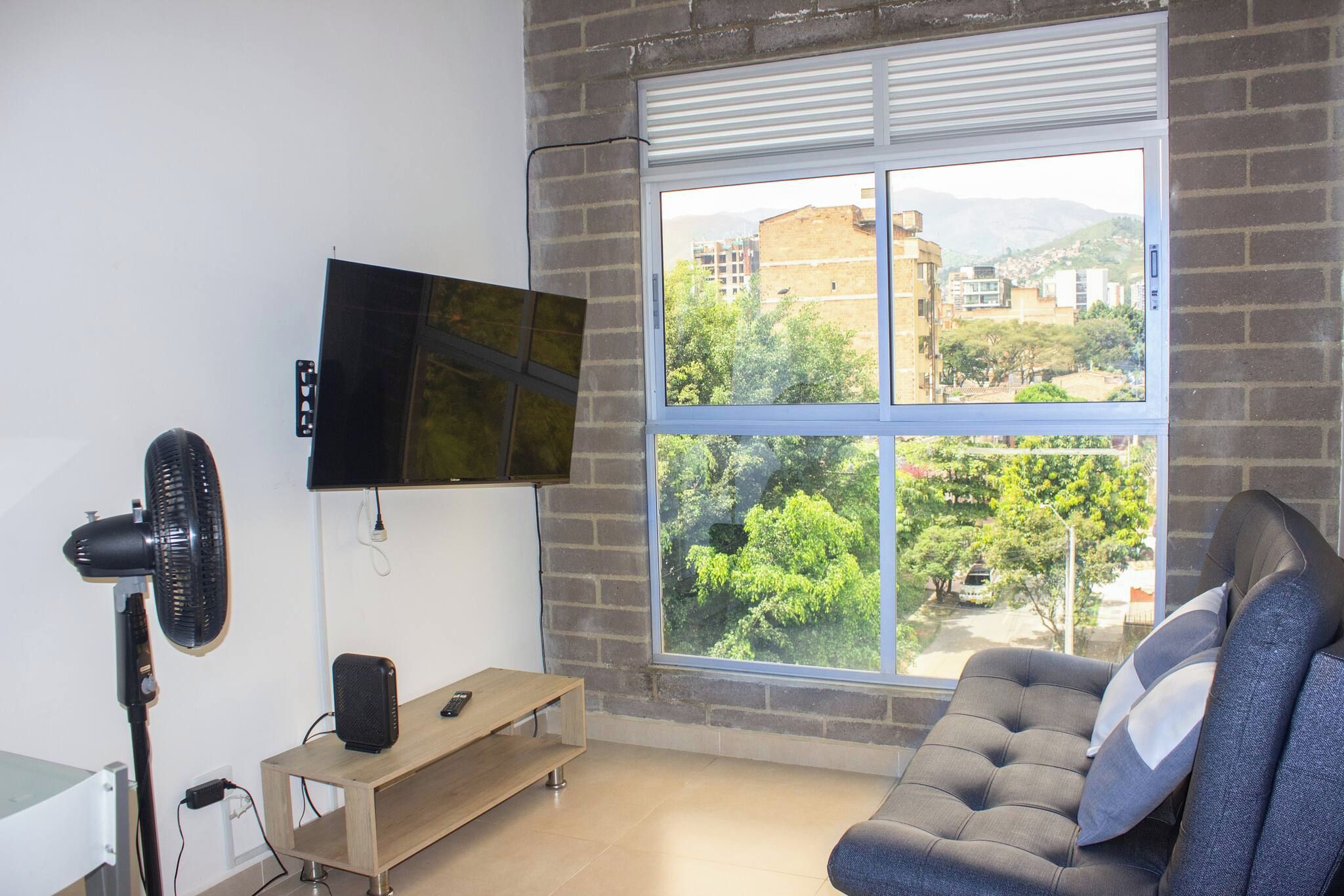 Una sala de estar con un televisor montado en la pared, un ventilador de pie, un sofá y una ventana con vistas a edificios y árboles.