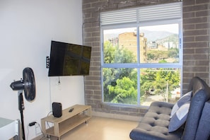 A living room with a TV mounted on the wall, a standing fan, a sofa, and a window with a view of buildings and trees. - 51 Close to Laureles, Metro and Restaurants (Medellín)