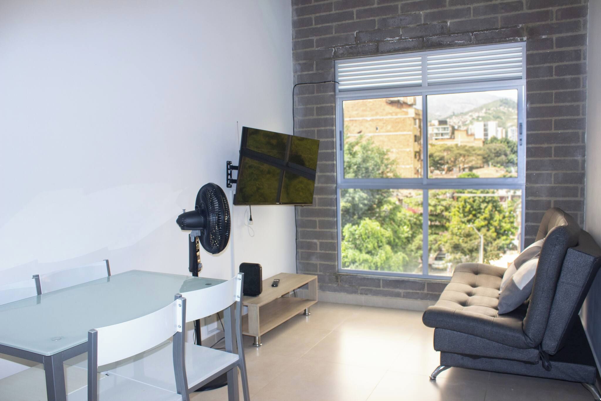 Una habitación moderna con una mesa de vidrio, un sofá, un televisor montado en la pared y un ventanal con vistas a zonas verdes y edificios.