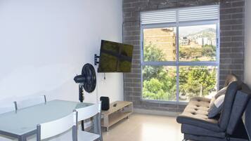 Una habitación moderna con una mesa de vidrio, un sofá, un televisor montado en la pared y un ventanal con vistas a zonas verdes y edificios.