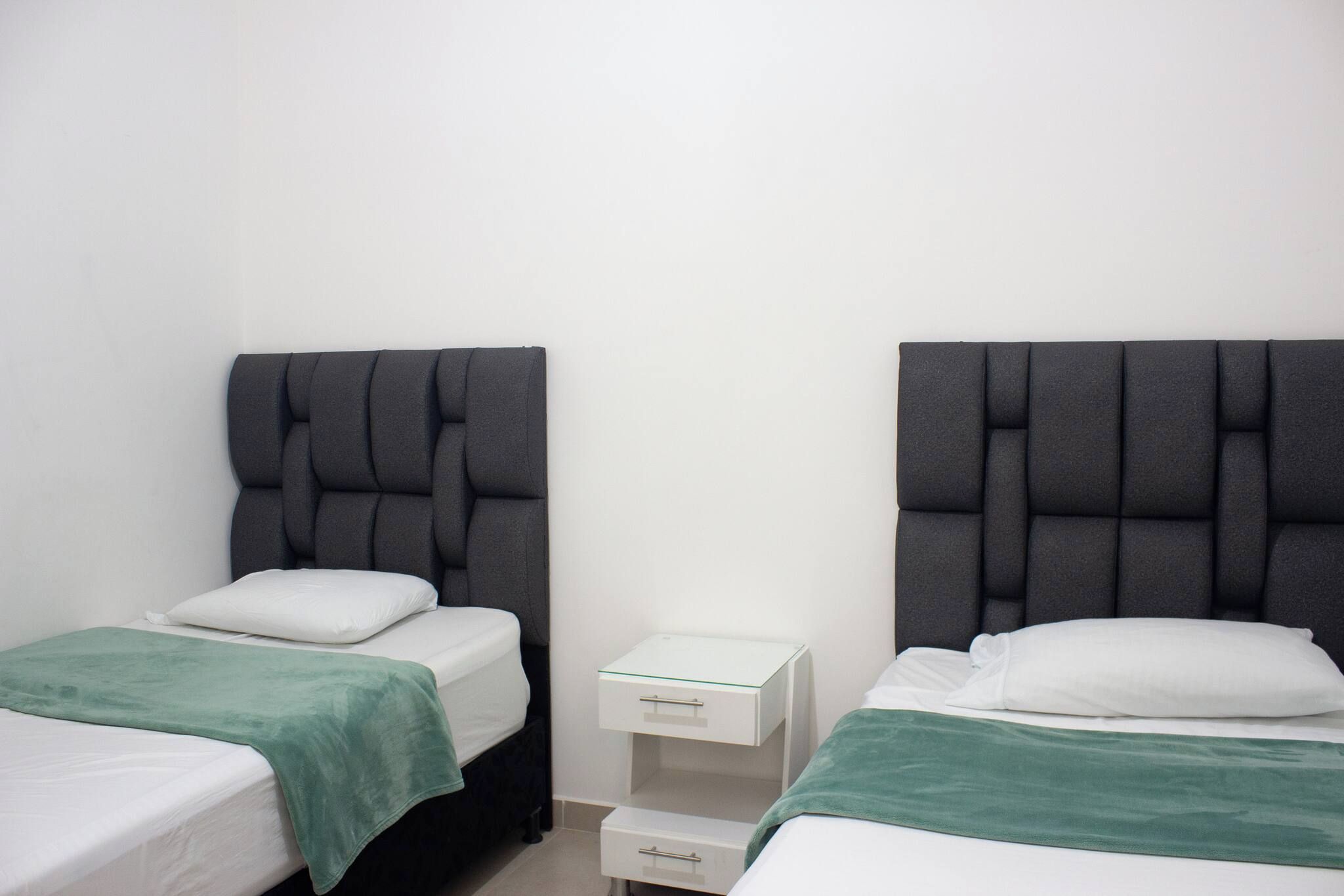 Una habitación con dos camas, cada una con cabecero gris y ropa de cama blanca. Hay una mesita de noche pequeña y blanca con cajones entre las camas.