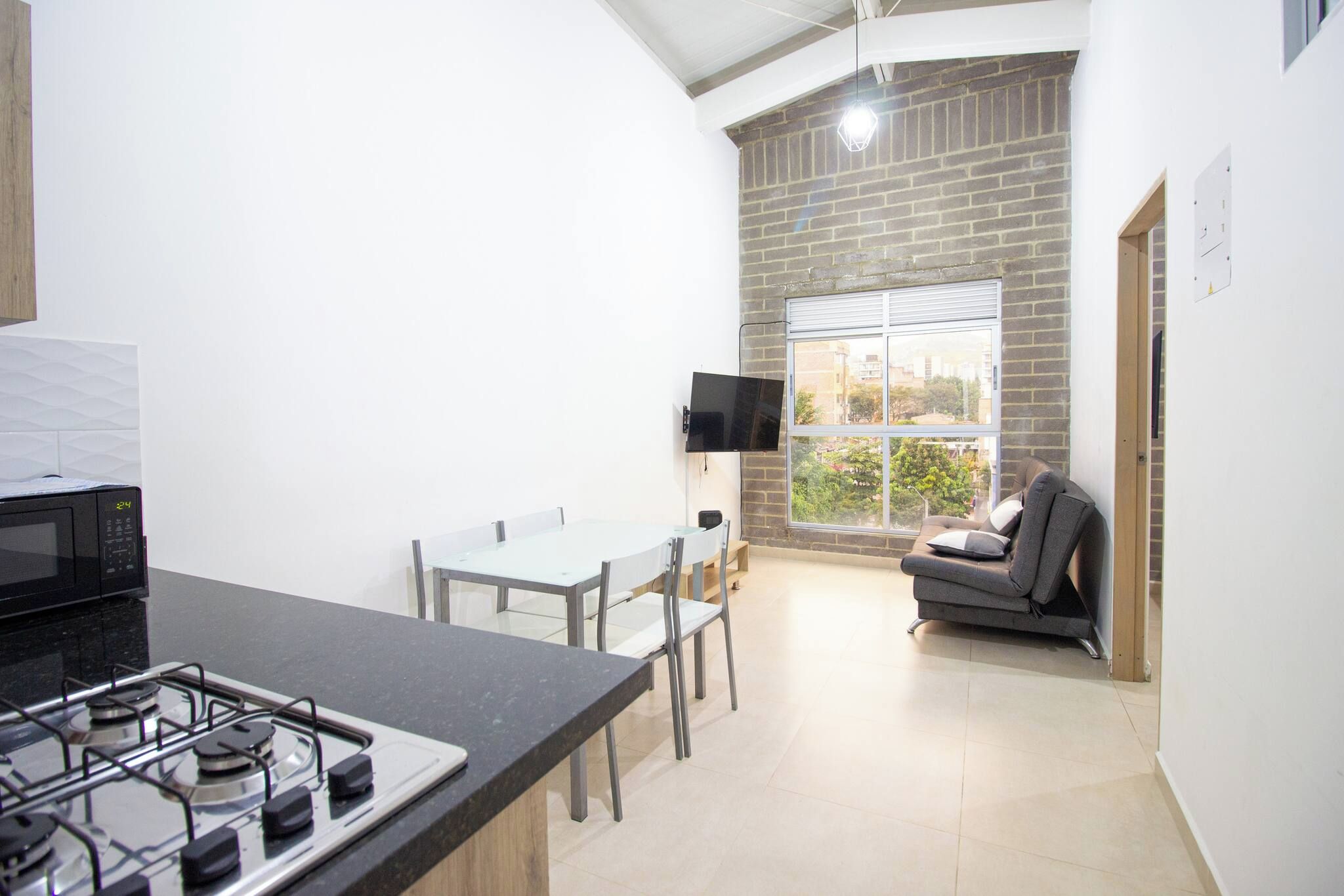 Cocina compacta con estufa a gas, microondas, mesa de comedor con sillas, televisor de pantalla plana montado en la pared y sofá en un rincón con vistas al exterior.