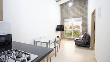 Cocina compacta con estufa a gas, microondas, mesa de comedor con sillas, televisor de pantalla plana montado en la pared y sofá en un rincón con vistas al exterior.