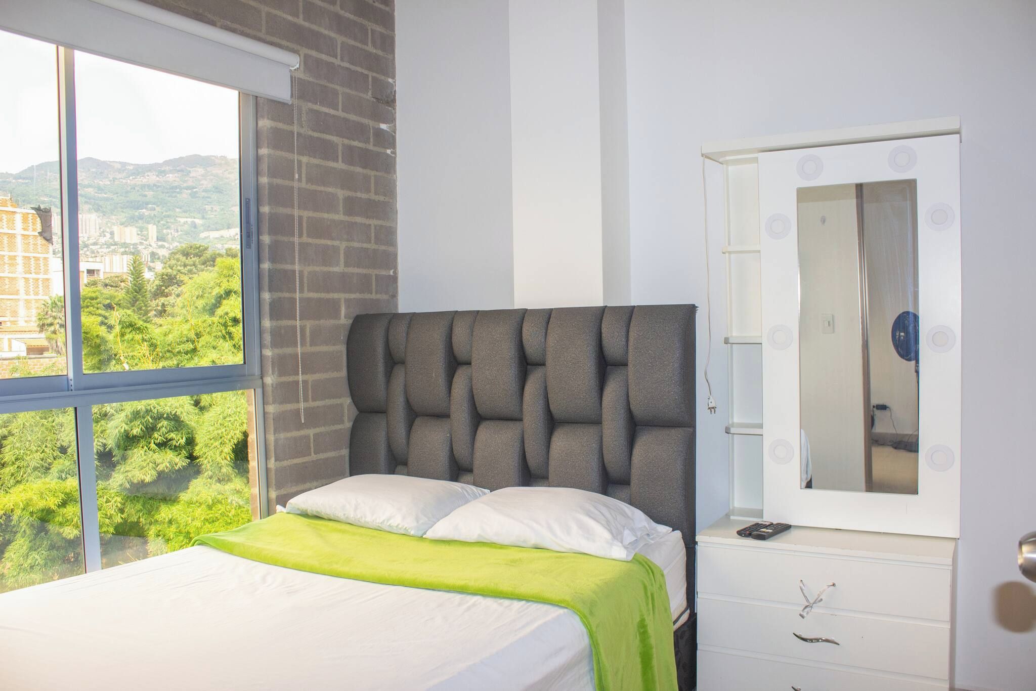 A bedroom with a bed, a window with a view of greenery, a brick wall, and a white dresser with a mirror.