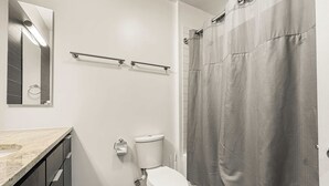 A bathroom with a toilet, a sink, a mirror, a shower curtain, and a trash bin. - Compact Studio for a Relaxed Stay (Chicago)