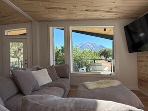 A modern living room with a grey sofa, a fluffy throw, and a TV mounted on the wall. Large windows offer a view of a mountain and outdoor furniture. - Sopris Mountain Retreat w/ Pool & Mountain Views (Carbondale)