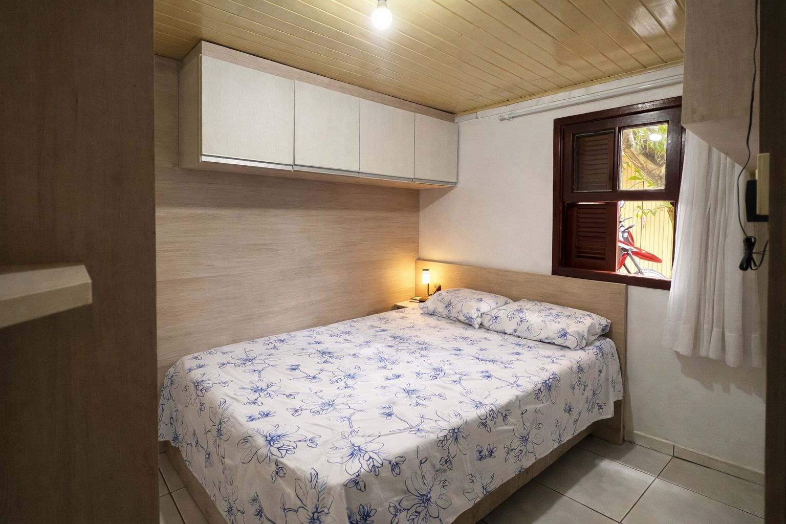 A small bedroom with a single bed, a nightstand, a window with a view of a bicycle, and a wooden ceiling.