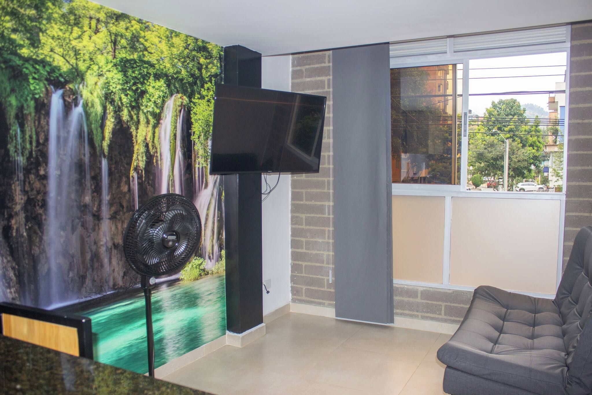 Una sala de estar con un mural de cascada, un televisor de pantalla plana, un ventilador de pie y un sofá gris.