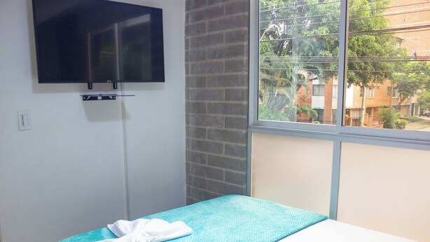Quarto com uma cama de solteiro, TV de tela plana na parede e janela com vista para edifícios e árvores.