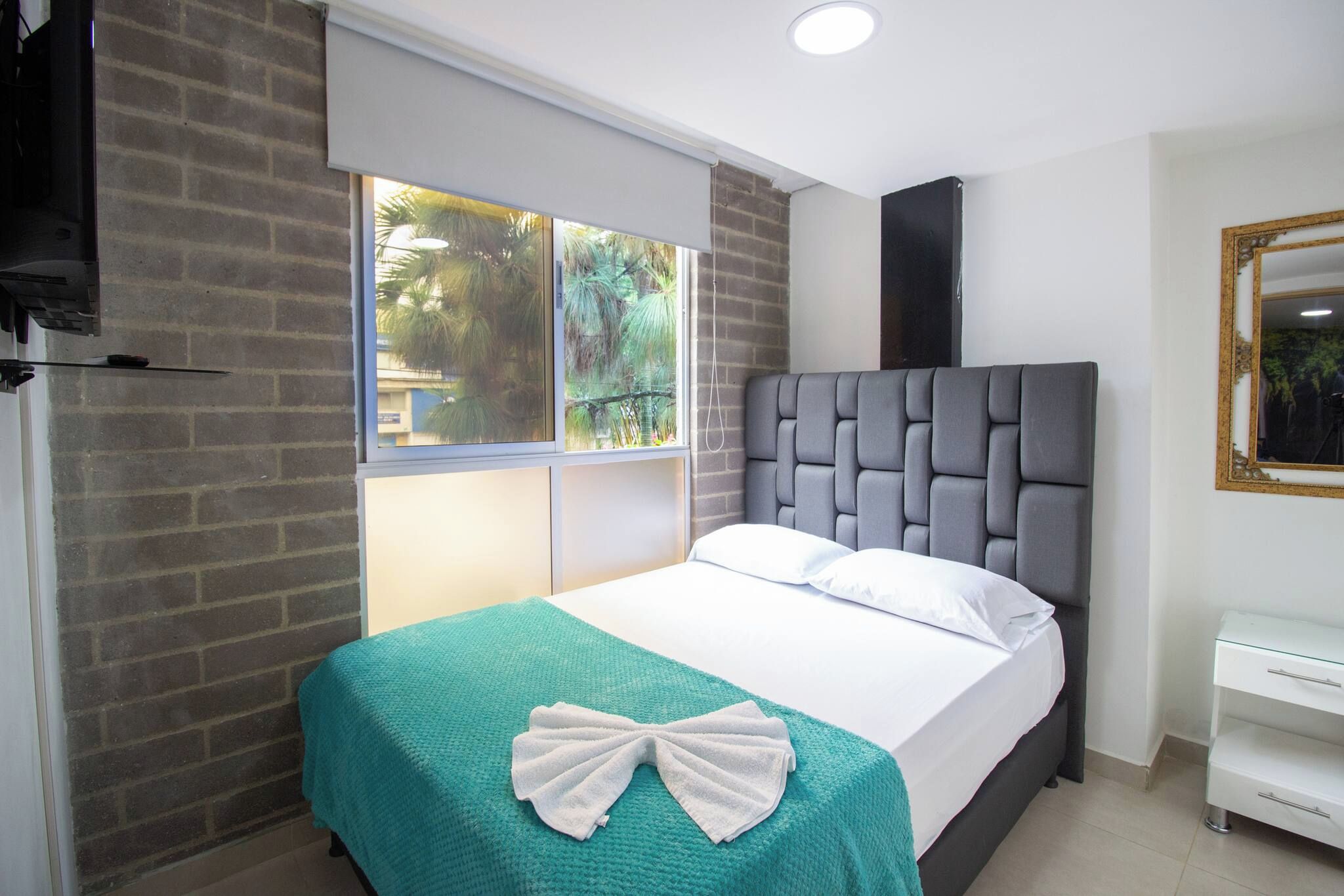 Habitación de hotel con cama, ventana con vista a follaje, mesita de noche blanca y espejo.