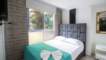Habitación de hotel con cama, ventana con vista a follaje, mesita de noche blanca y espejo.