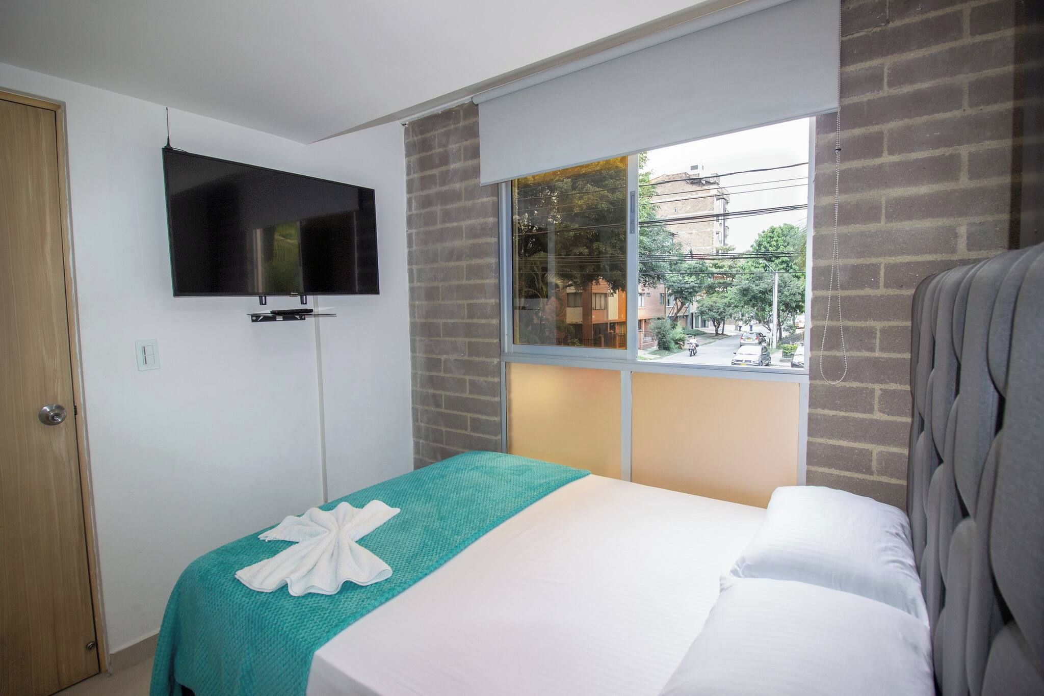 Habitación de hotel con cama, televisión montada en la pared, ventana con vista a la calle y una puerta.