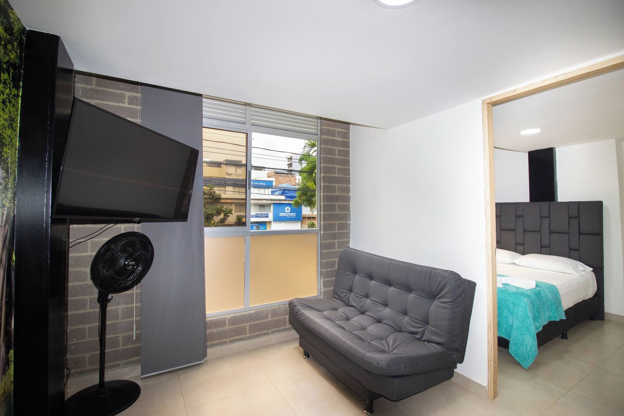 Un dormitorio con un sofá gris, una cama con colchón blanco, un televisor montado en la pared y una ventana con vistas a edificios.
