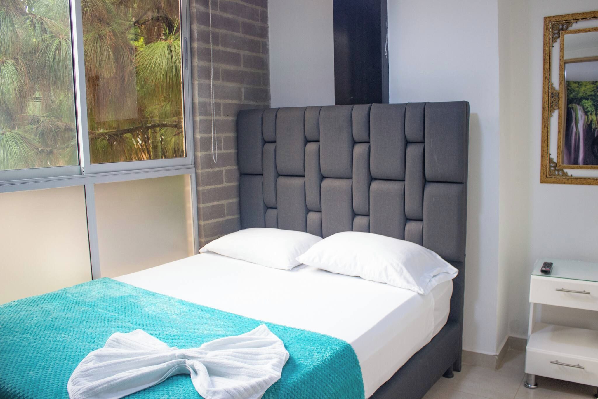 A neatly made bed with white linens and a teal blanket, a grey upholstered headboard, and a small bedside table with a remote control.
