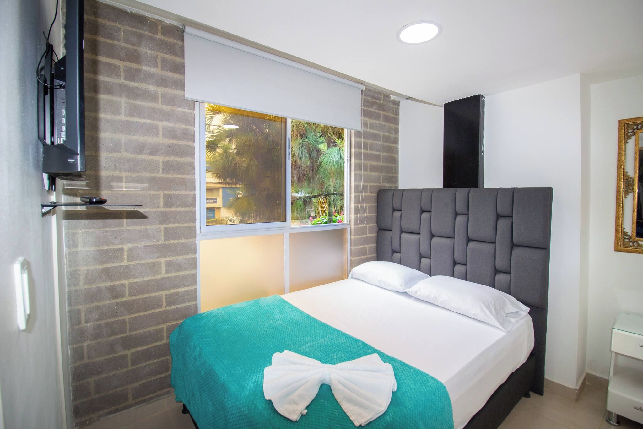 Habitación de hotel con cama, televisor, ventana con vista a follaje y televisor montado en la pared.