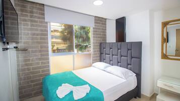 A hotel room with a bed, a window with a view of buildings and trees, a white bedside table, and a mirror with a decorative frame.