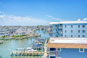 A marina with docked boats, a blue building with balconies, and a clear sky. - 3BR Bayfront | Boardwalk |Sunsets Balcony |Pool | Parking (Ocean City)