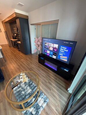 Een moderne woonkamer met een flatscreen televisie, houten vloeren, een glazen salontafel en een keuken in de achtergrond.