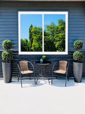 A patio with two chairs and a table, a window with a view of trees, and two potted plants.