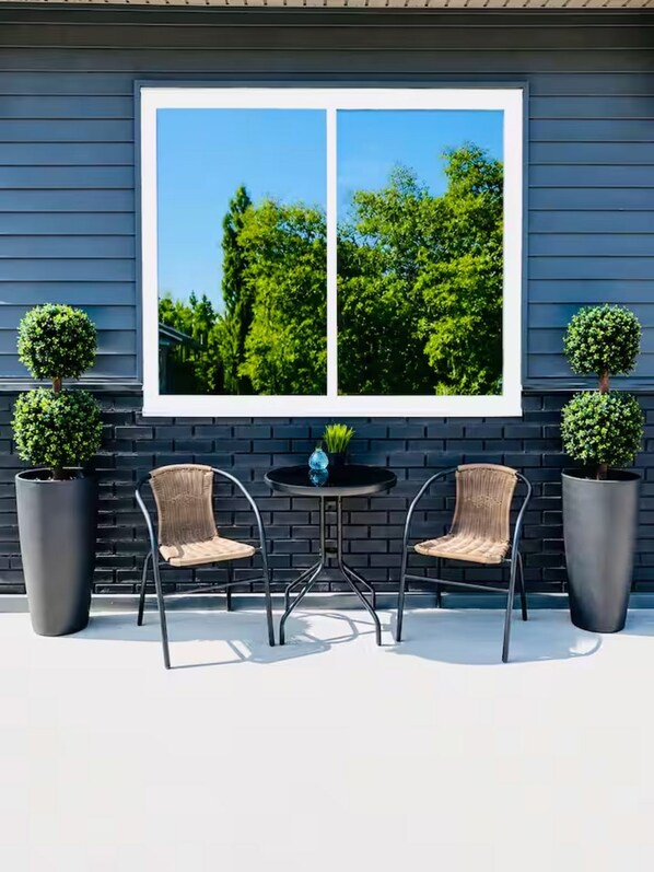 A patio with two chairs and a table, a window with a view of trees, and two potted plants.