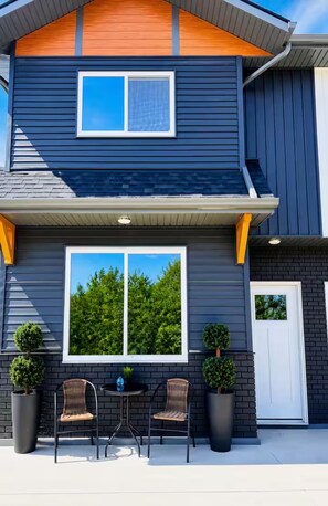 A two-story house with a dark exterior, a large window, and a small patio with chairs and potted plants.