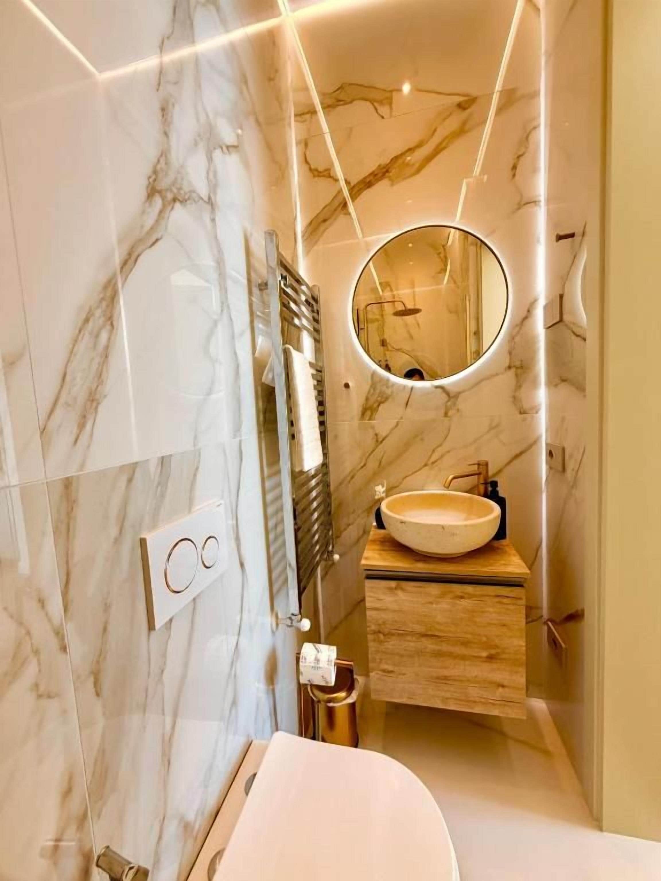 Un bagno moderno con una parete in marmo, uno specchio rotondo, un mobile contenitore in legno e un lavandino bianco.