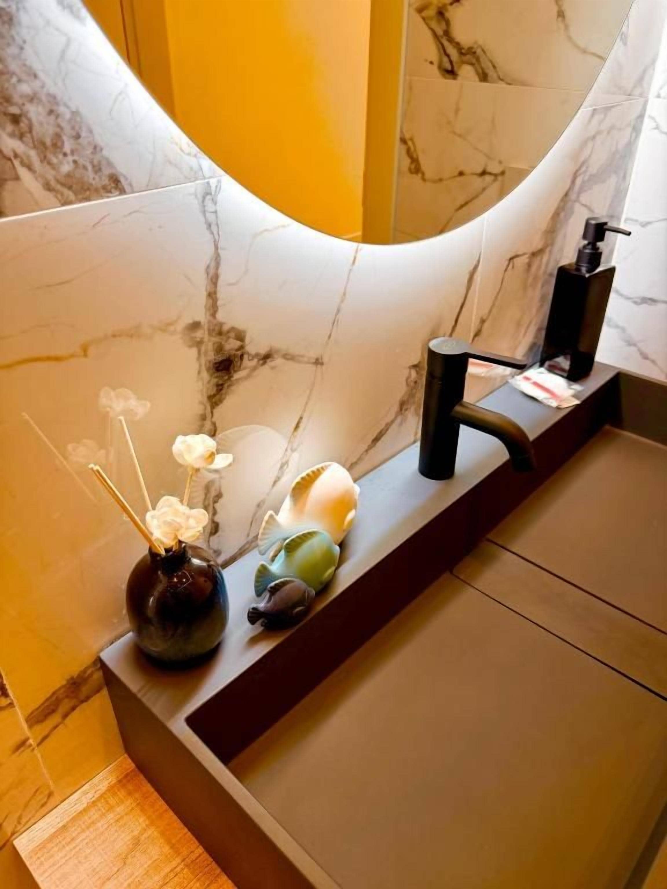 Un baño con pared de mármol, un lavamanos con grifo negro y elementos decorativos, incluyendo un jarrón con flores y conchas marinas.