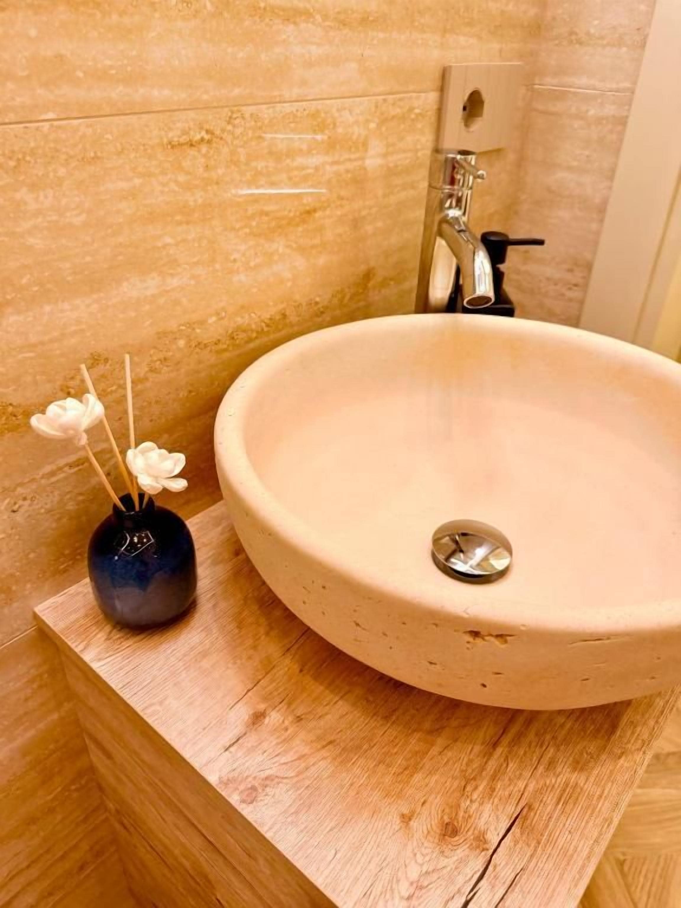 A bathroom with a round beige sink, a wooden countertop, and a small dark vase with flowers.