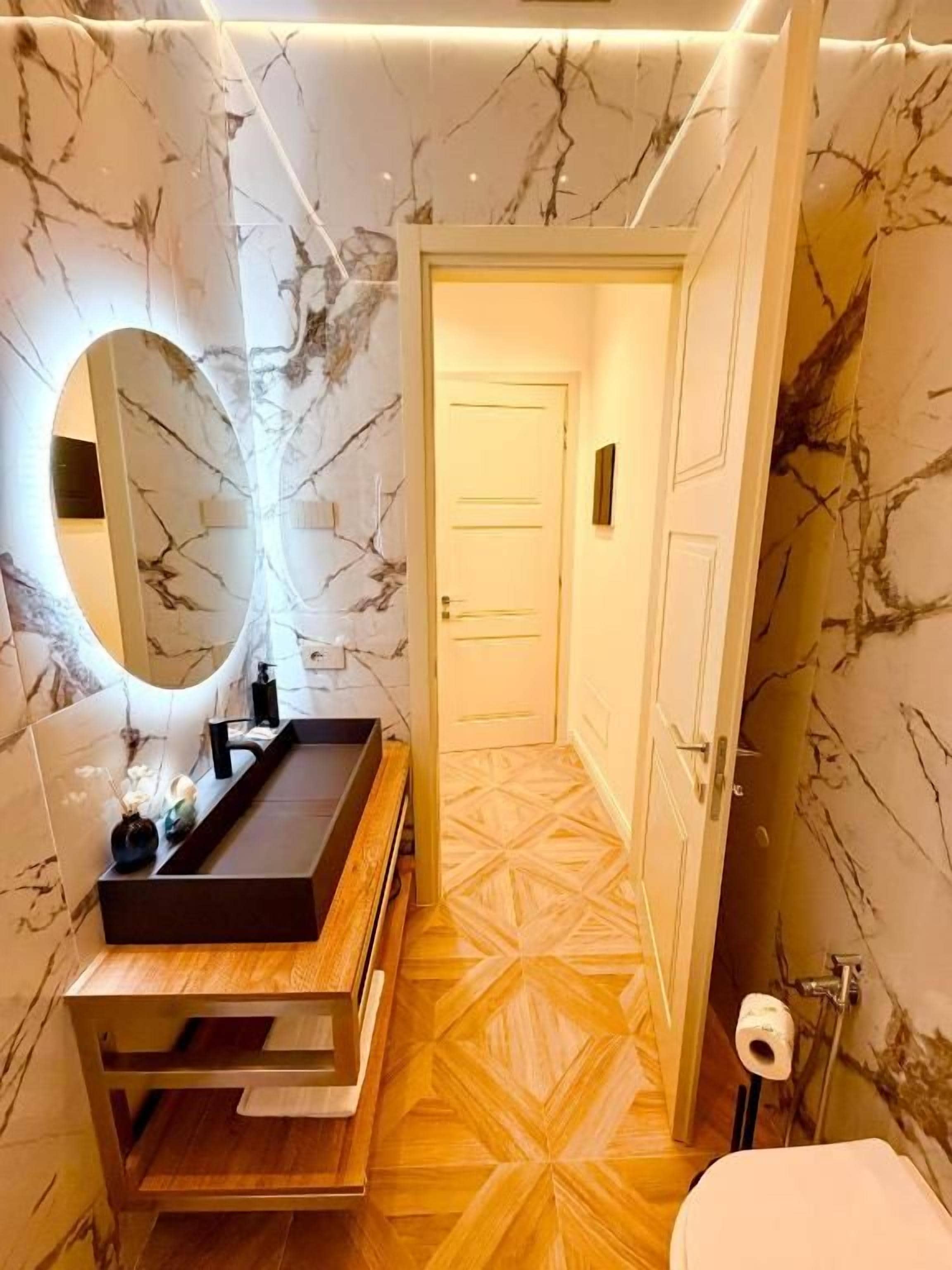 Une salle de bain avec un mur en marbre, un meuble-lavabo en bois, un miroir rond et un sol à motifs géométriques.