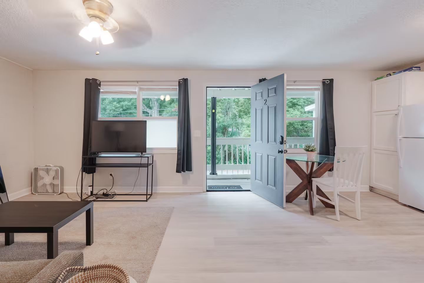 A modern living room with a flat-screen TV, a dining table with chairs, a white refrigerator, and a large window with curtains.
