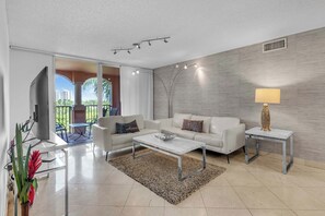 Ein modernes Wohnzimmer mit einem weißen Sofa, einem Couchtisch und einer Lampe. Blick auf einen Balkon mit Gartenmöbeln.