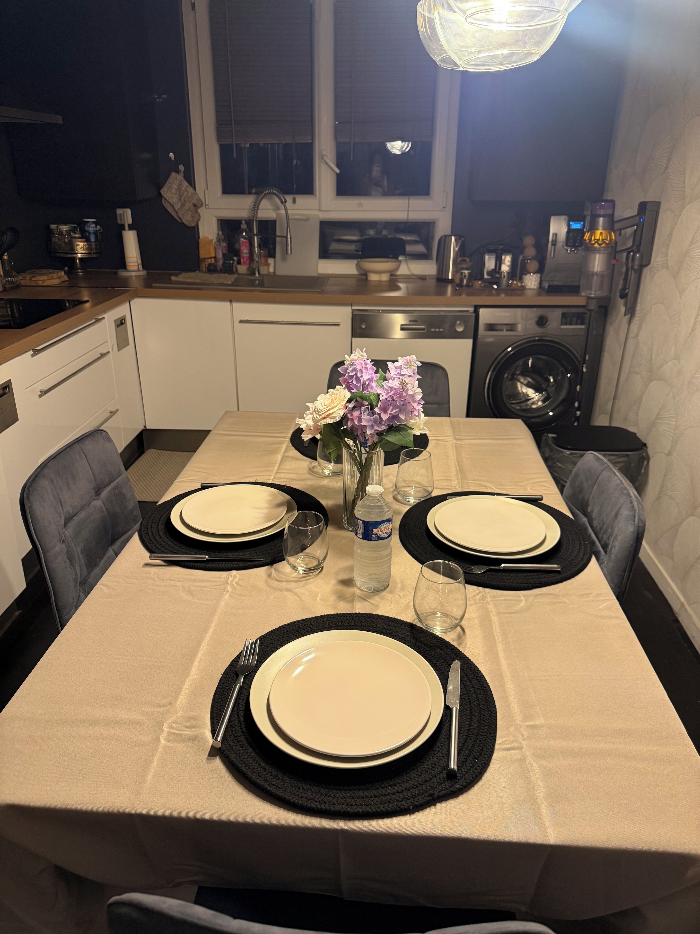 Une table à manger dressée avec des assiettes, des couverts et un vase de fleurs, dans une cuisine moderne équipée d’un lave-linge et de placards.