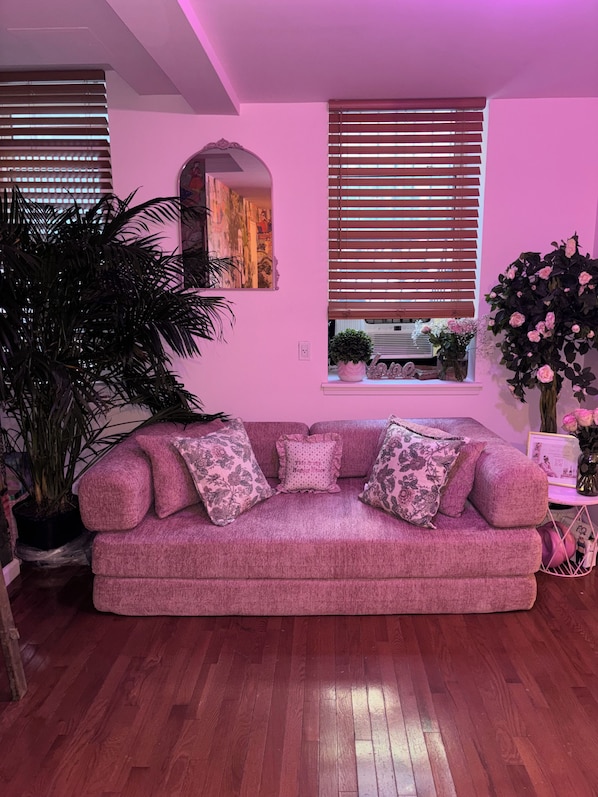 Un canapé rose agrémenté de coussins à motifs floraux, dans une pièce au plancher en bois, avec une grande plante en pot et une fenêtre équipée de stores.