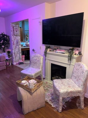 Un salon avec une cheminée, deux fauteuils à motifs floraux, un téléviseur et un plateau contenant des objets décoratifs.