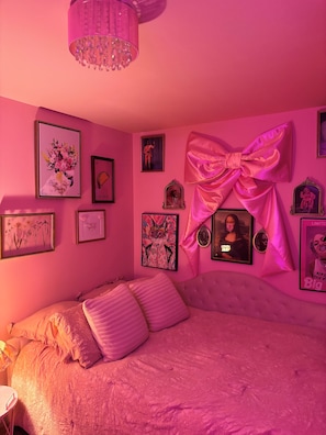 Une chambre à coucher décorée dans des tons de rose, avec un grand lit, un lustre et divers tableaux encadrés accrochés aux murs.