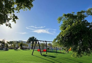 Ein Park mit Schaukeln, grüner Wiese, Bäumen und Spielgeräten.