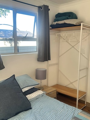 A bedroom with a bunk bed, a bedside table with a lamp, a window with curtains, and a white wardrobe with hangers.