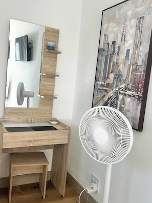 A wooden desk with a mirror, a fan, and a cityscape painting on the wall.