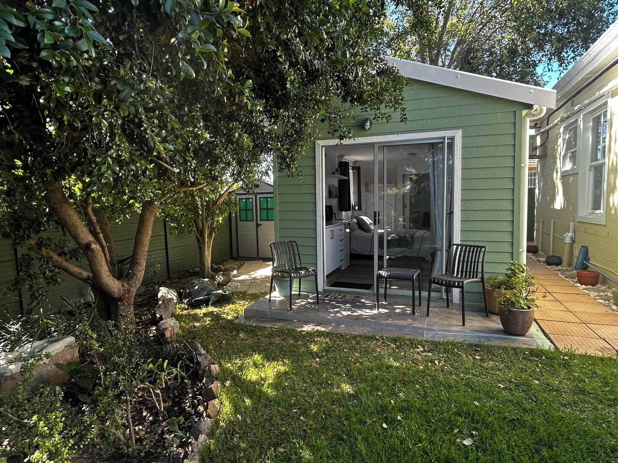 Uma casa pequena e verde com uma porta de correr de vidro que dá para um quarto. Há duas cadeiras do lado de fora e um vaso de plantas no alpendre.