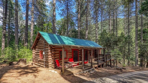 A log cabin with a green roof, a porch with red chairs, and a dirt path leading to it. - Fir - Rustic Log Cabin (Sierra City)