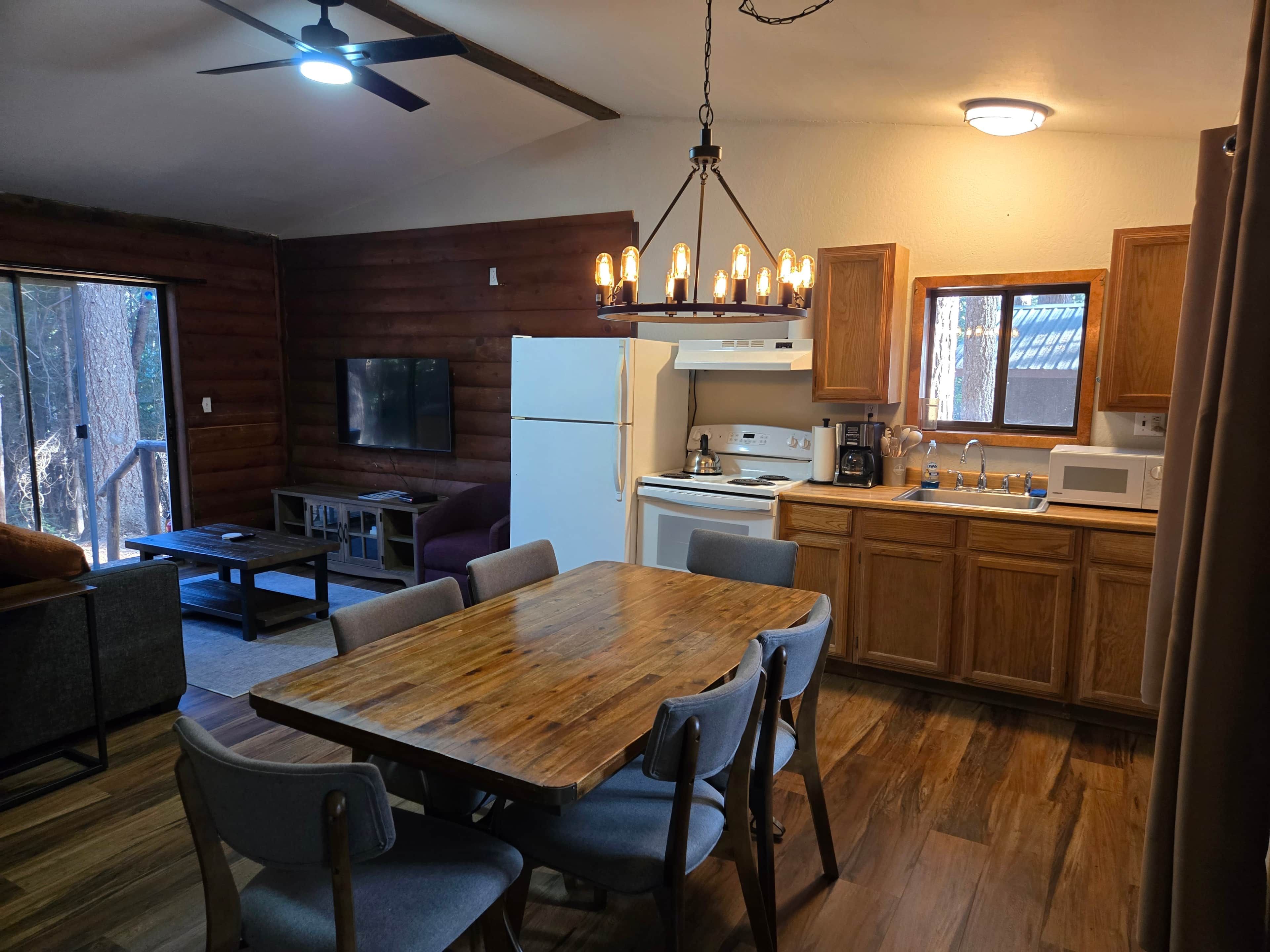 L’intérieur chaleureux d’un chalet avec une table à manger en bois, des chaises, une cuisine équipée d’une cuisinière et d’un réfrigérateur, et un téléviseur fixé au mur.
