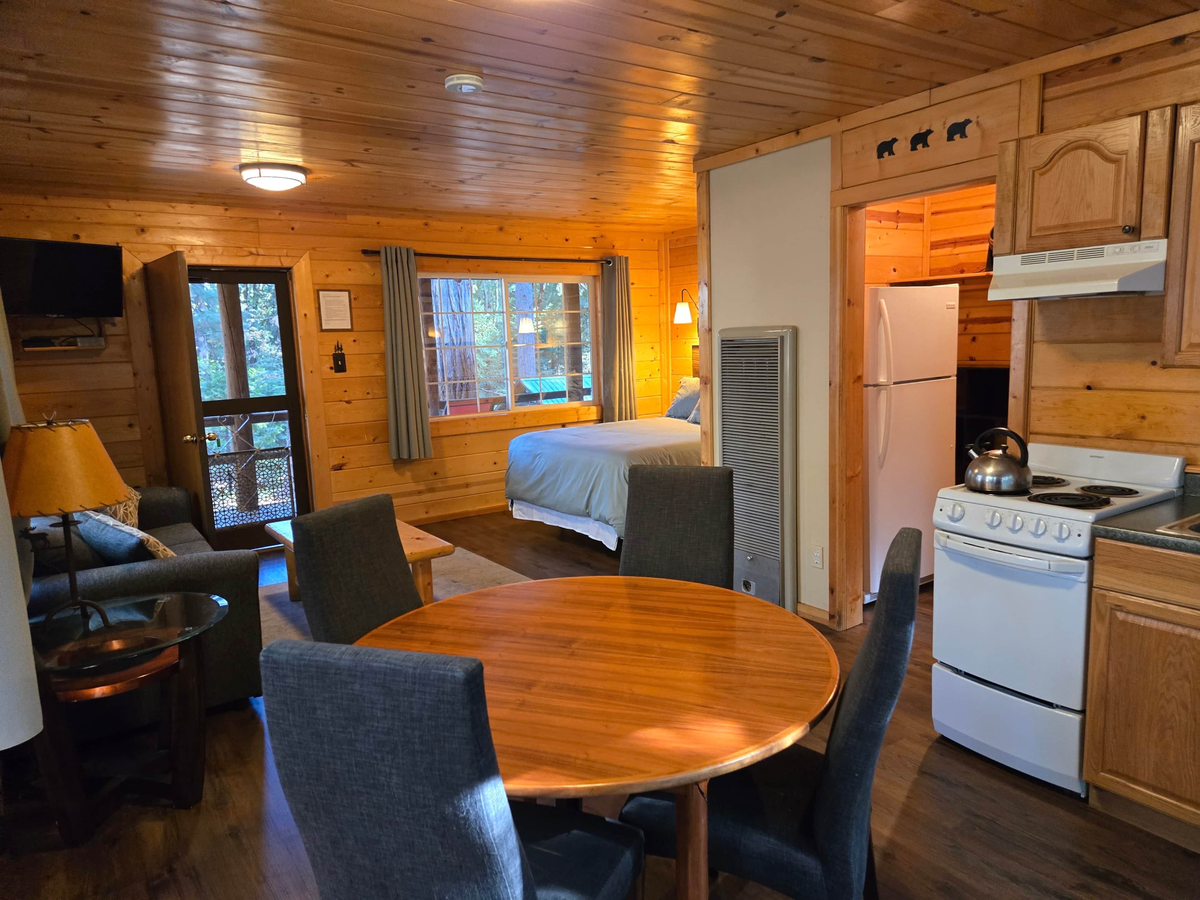 Interior de una cabaña con una cama, mesa de comedor, sillas, una cocina con estufa y un televisor.